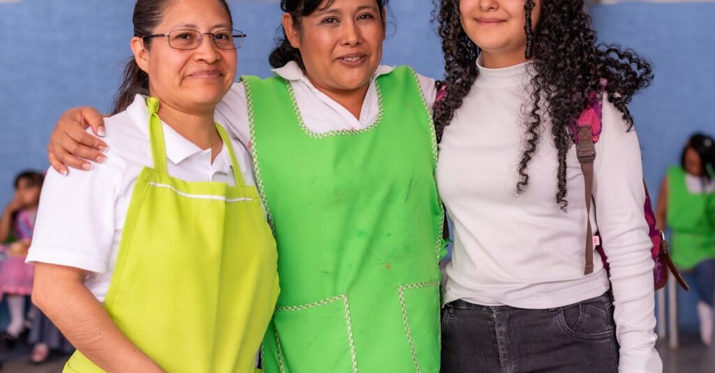 Three women indoors, smiling and wearing green aprons, showing togetherness.